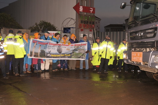 Veterans For Peace delegation stands with banner while giant construction vehicles leave the site. Photo by ELLEN DAVIDSON